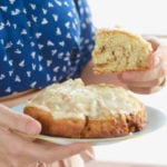 A hand holding a plate with a Giant Cinnamon Roll on it, with a slice in the other hand.