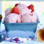 A tub of Homemade Strawberry Gelato, topped with strawberries and flanked by a scoop and cones.