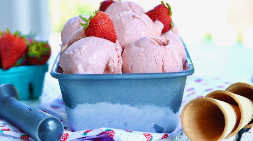 A tub of Homemade Strawberry Gelato, topped with strawberries and flanked by a scoop and cones.