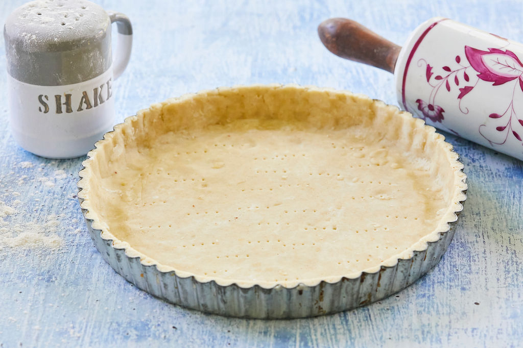 A gluten-free pie crust, ready for filling, flanked by a rolling pin and shaker.