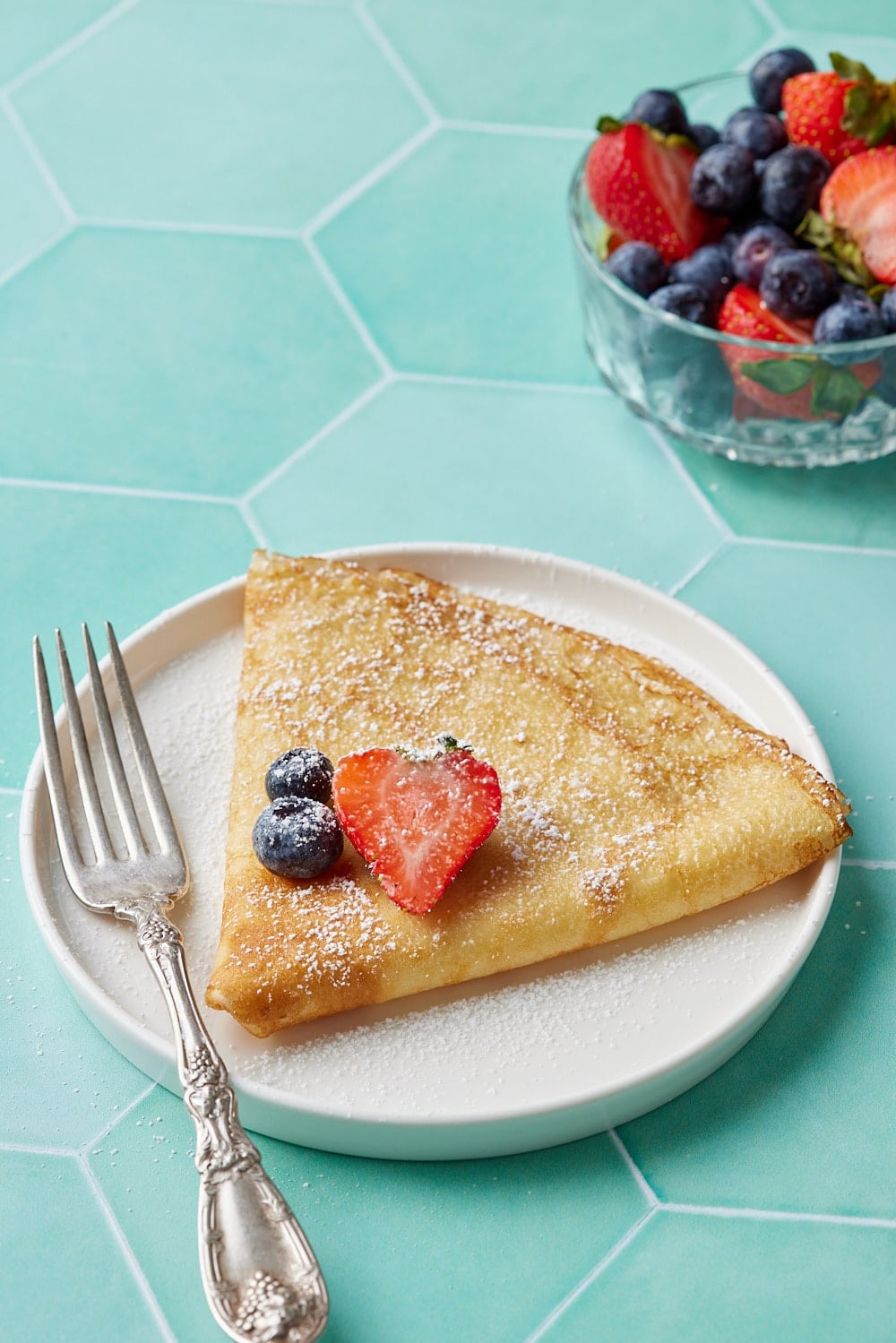 A piece of crepe folded into a triangle served with two blueberries and one slice of strawberry on top. 