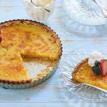 Lemon tart with golden crust and filling topped with fresh berries. A slice is cut and served on the side.