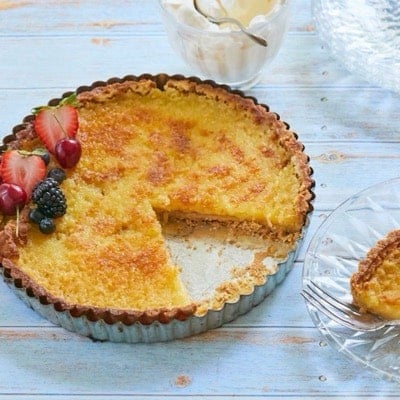 Lemon tart with golden crust and filling topped with fresh berries. A slice is cut and served on the side.