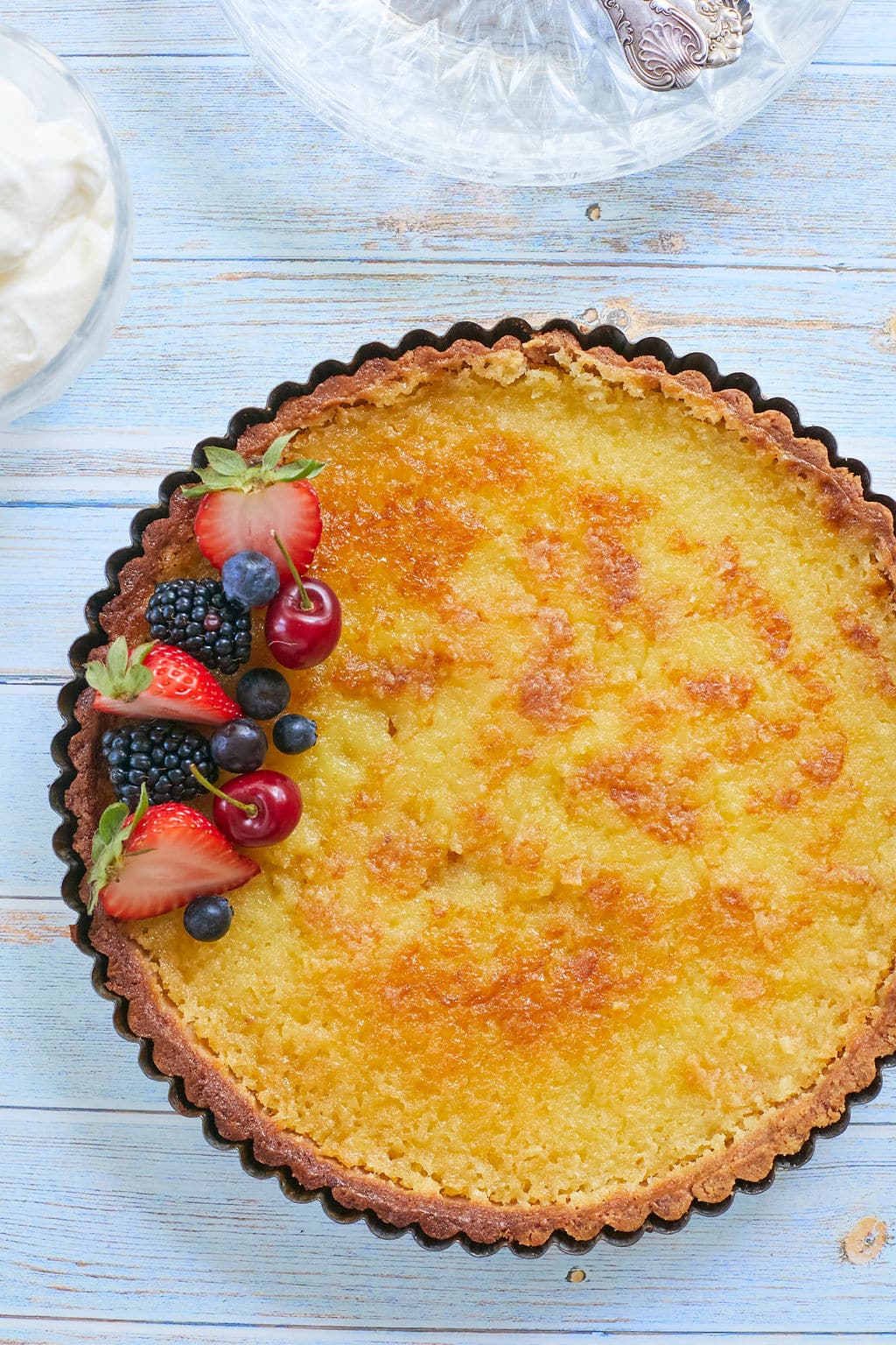 Lemon Tart7 Top down view of a golden lemon tart with fresh strawberries and blueberries. Served with softly whipped cream.