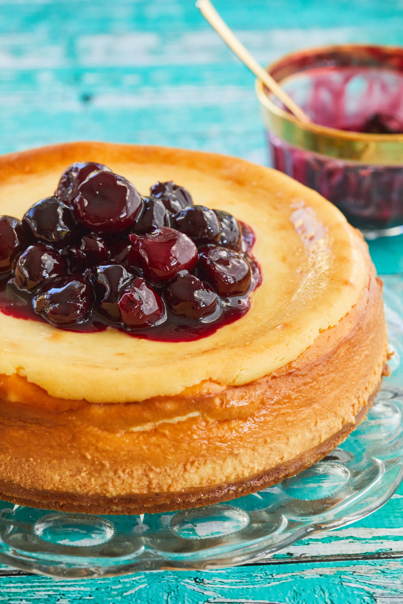 A New York cheesecake on the glass plate is perfectly baked golden, creamy, dense and sweet. It's topped with a big dollop of Candied Cherries (Glacé Cherries). A small glass bowl of Candied Cherries (Glacé Cherries) is on the side. 