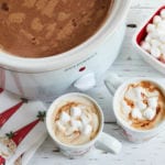 A slow cooker full of hot chocolate next to two mugs and a bowl of marshmallows.