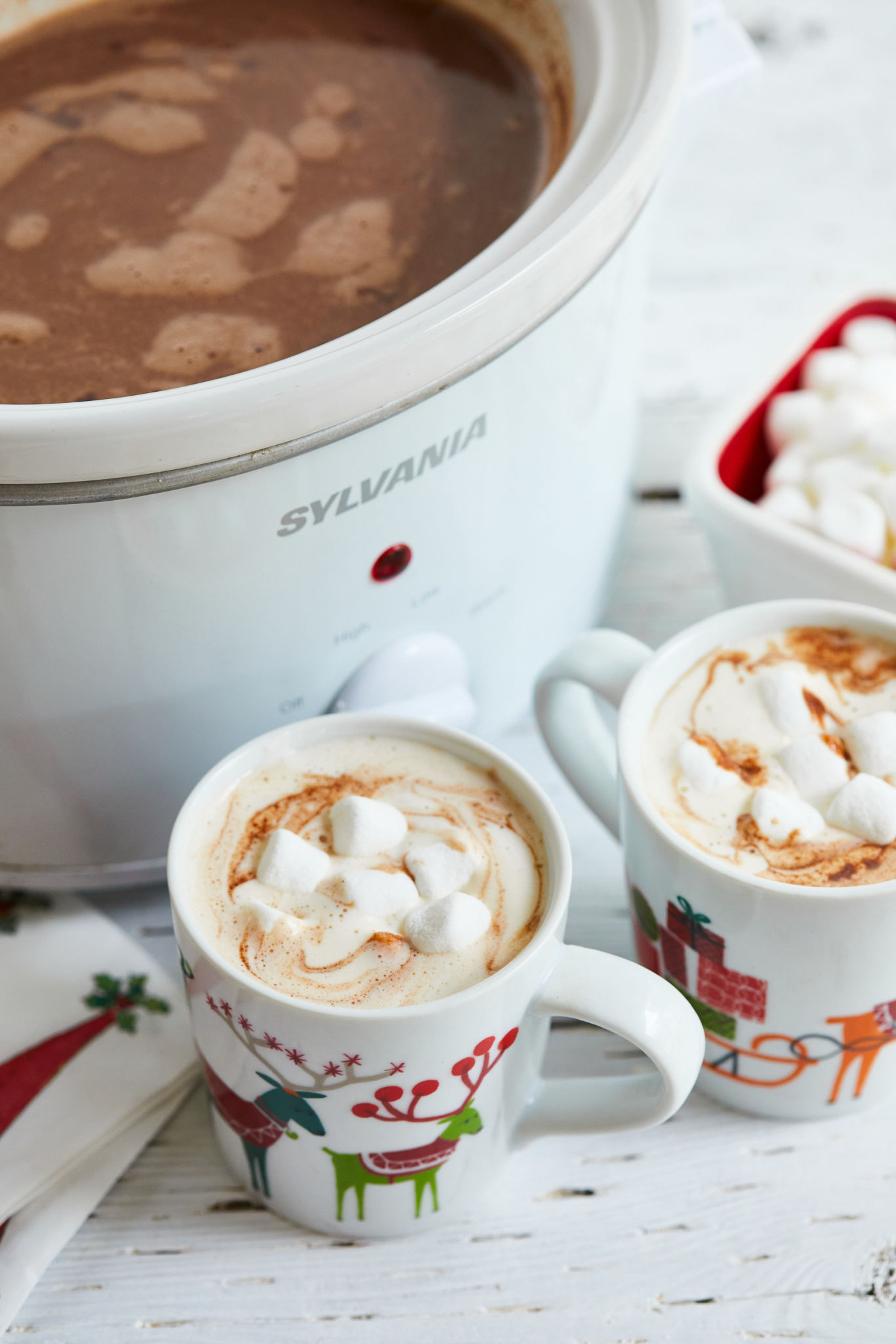 Two mugs of hot chocolate next to a slow cooker full of hot cocoa, and marshmallows.