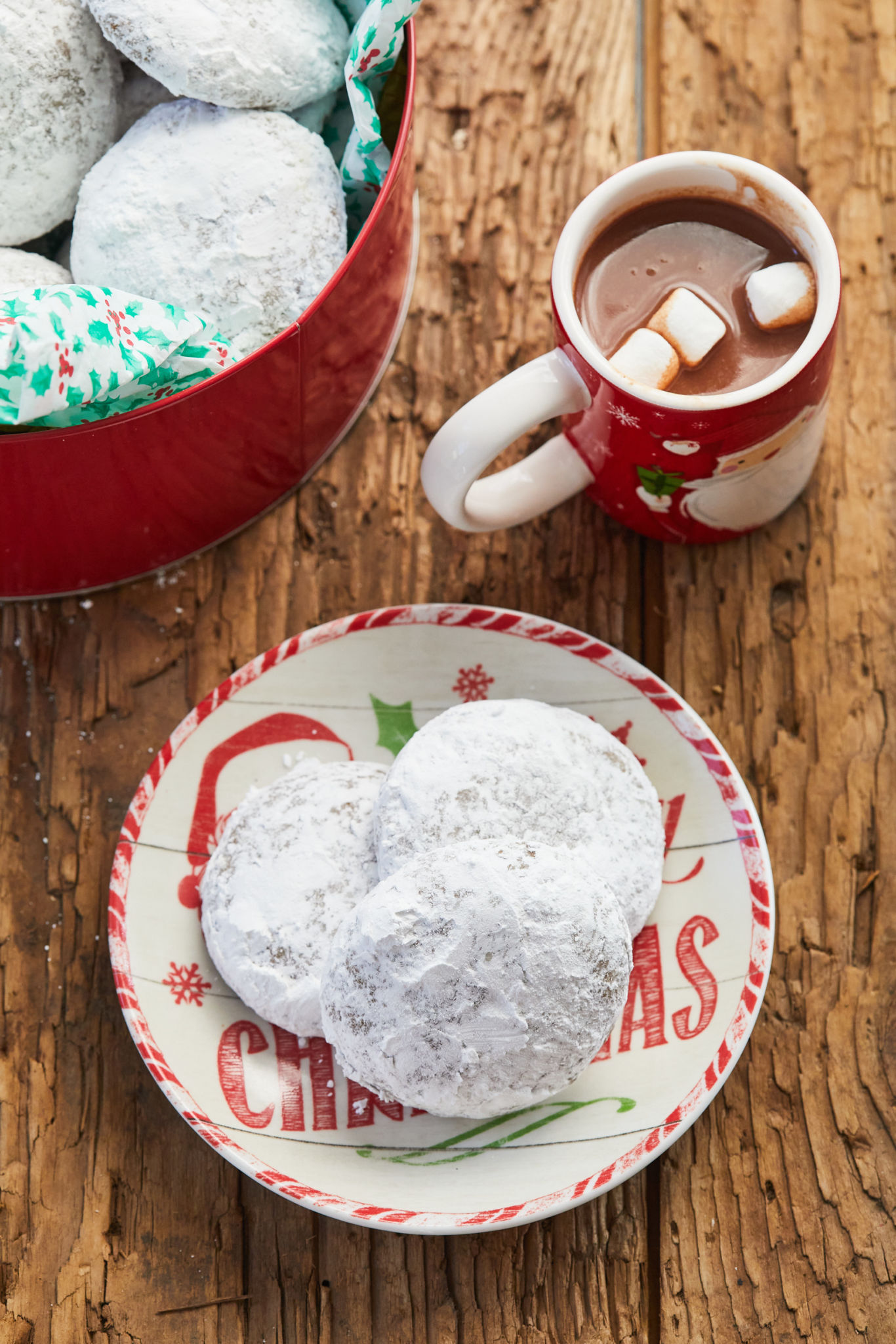 Homemade Spiced Pfeffernusse cookies, coated in powdered sugar, are served on a Christmas plate next to a bowl full of cookies and a Santa mug filled with hot chocolate with marshmallows. 