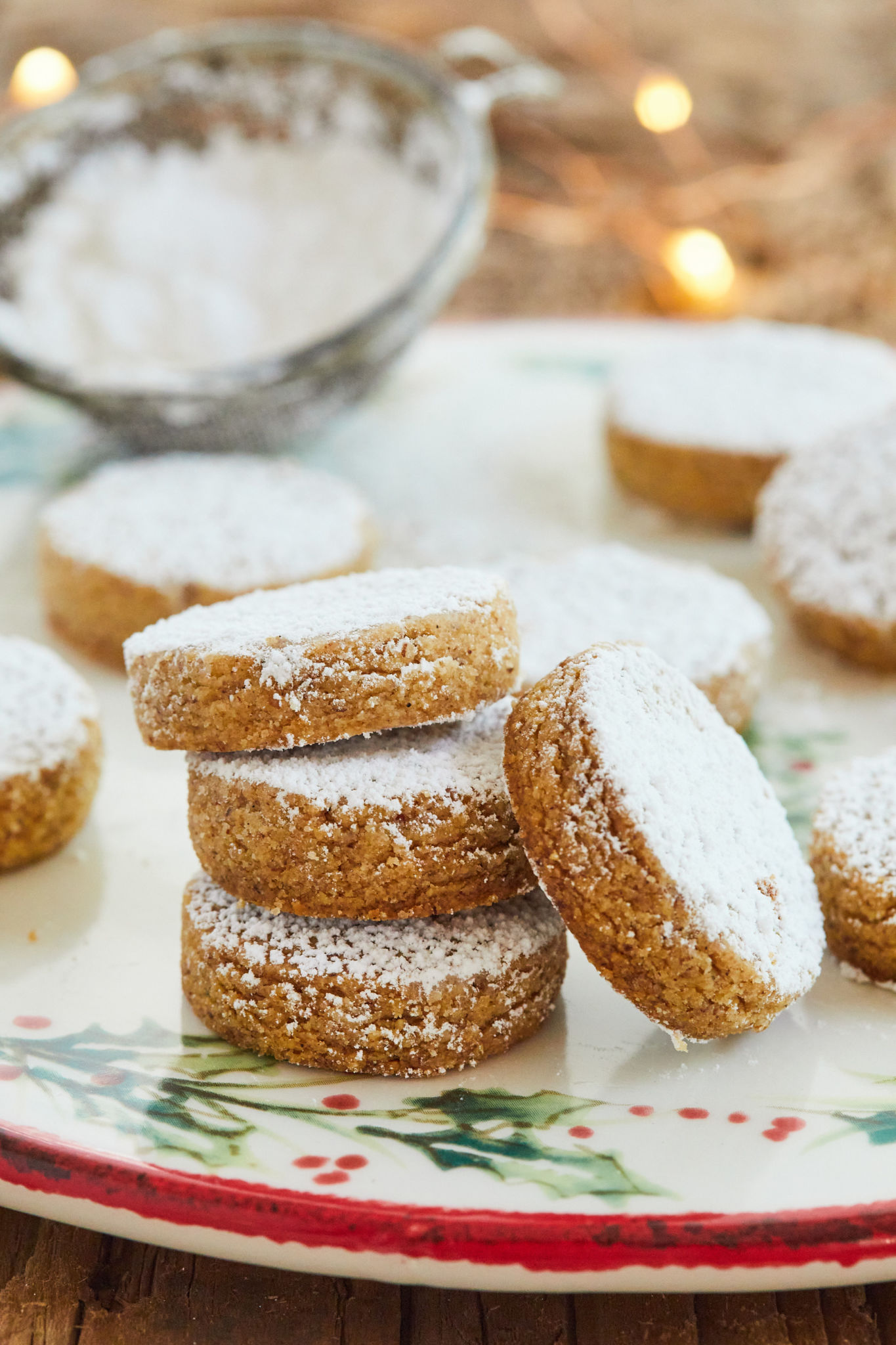 Polvorones, or Spanish almond cookies, are served on a plate decorated with holly berries. The cookies are slightly thick and covered in powdered sugar. 