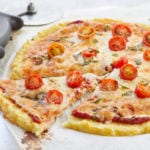 An entire cauliflower pizza, baked and topped with toppings, with one slice cut.