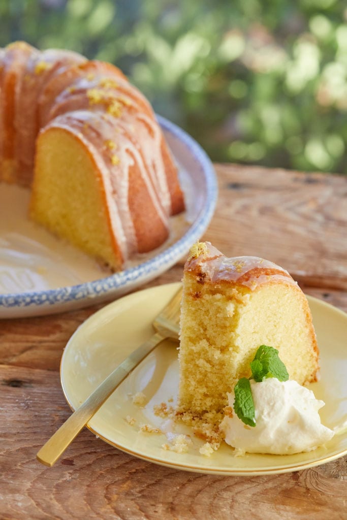 Moist, bright yellow Lemon Pound Cake is glazed and displayed on a big round platter. A slice is served on a dessert plate with whipped cream.