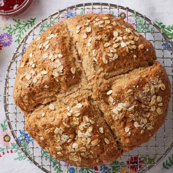 A big loaf of rustic, golden Irish soda bread with a cross scoring and topped with rolled oats.