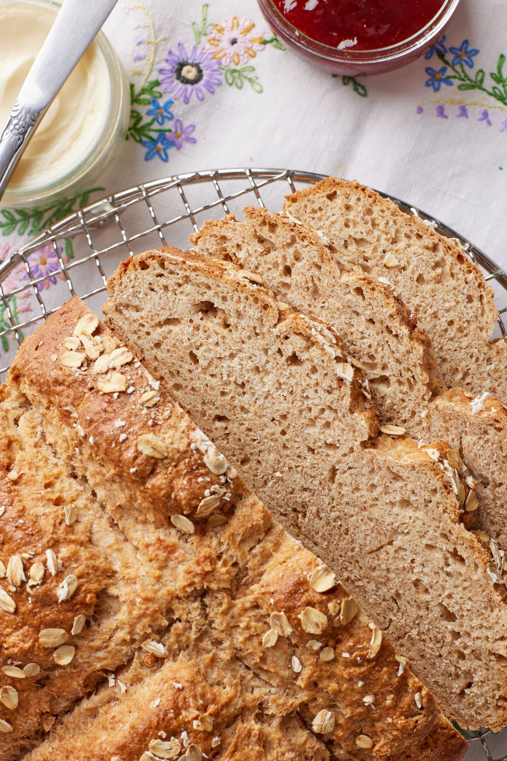 Half loaf and some slices of Irish Brown Soda Bread are served in a wire cooling rack, A jar of jam and a jar of butter are served in the side.