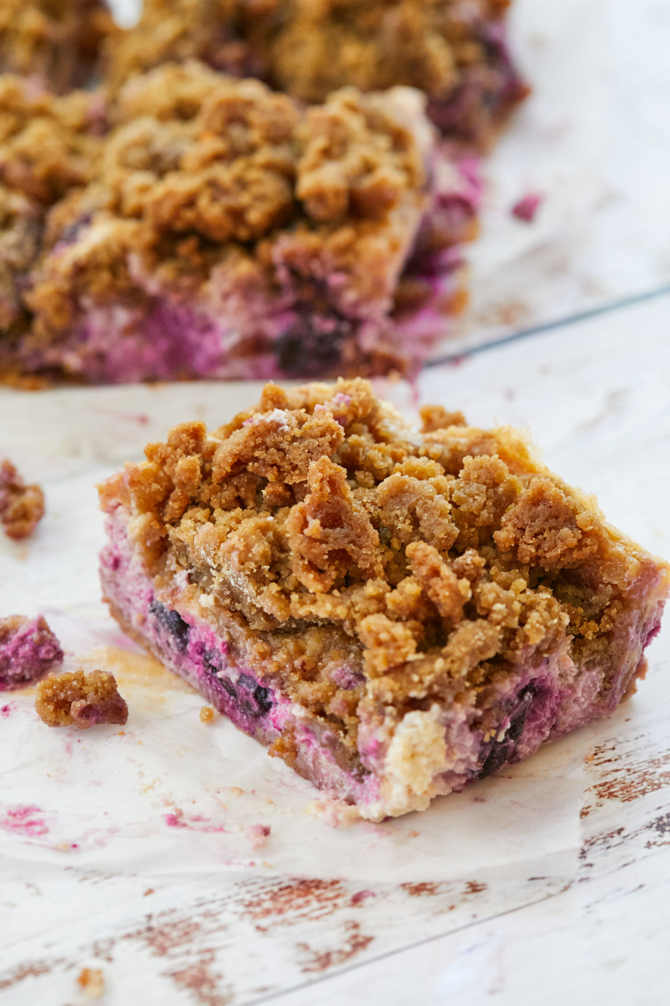 A square of Lemon and Blueberry cheesecake is cut off the whole cake that's behind it. It has gorgeous 3 ayers with crumbly buttery graham cracker crust at the bottom, sweet, tangy and creamy filling in the middle loaded with fruity and floral juicy blueberries, topped with crispy indulgent crumble topping.