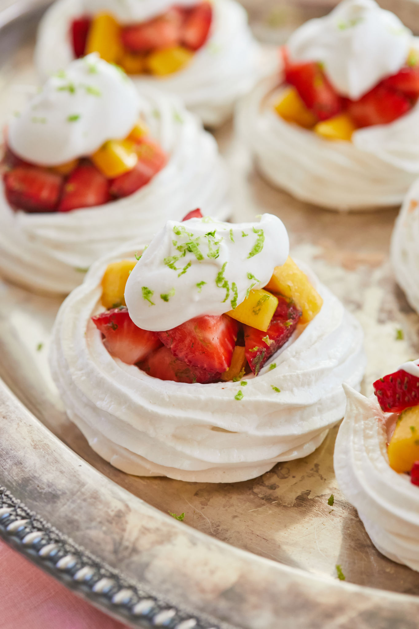 Stunning, heavenly meringue nests are placed on a silver platter. They are white layers of crispy billowy meringue loaded with irresistible tropical red strawberries and vibrant yellow mangoes, and adorned with velvety coconut cream, and green refreshing lime zest. 