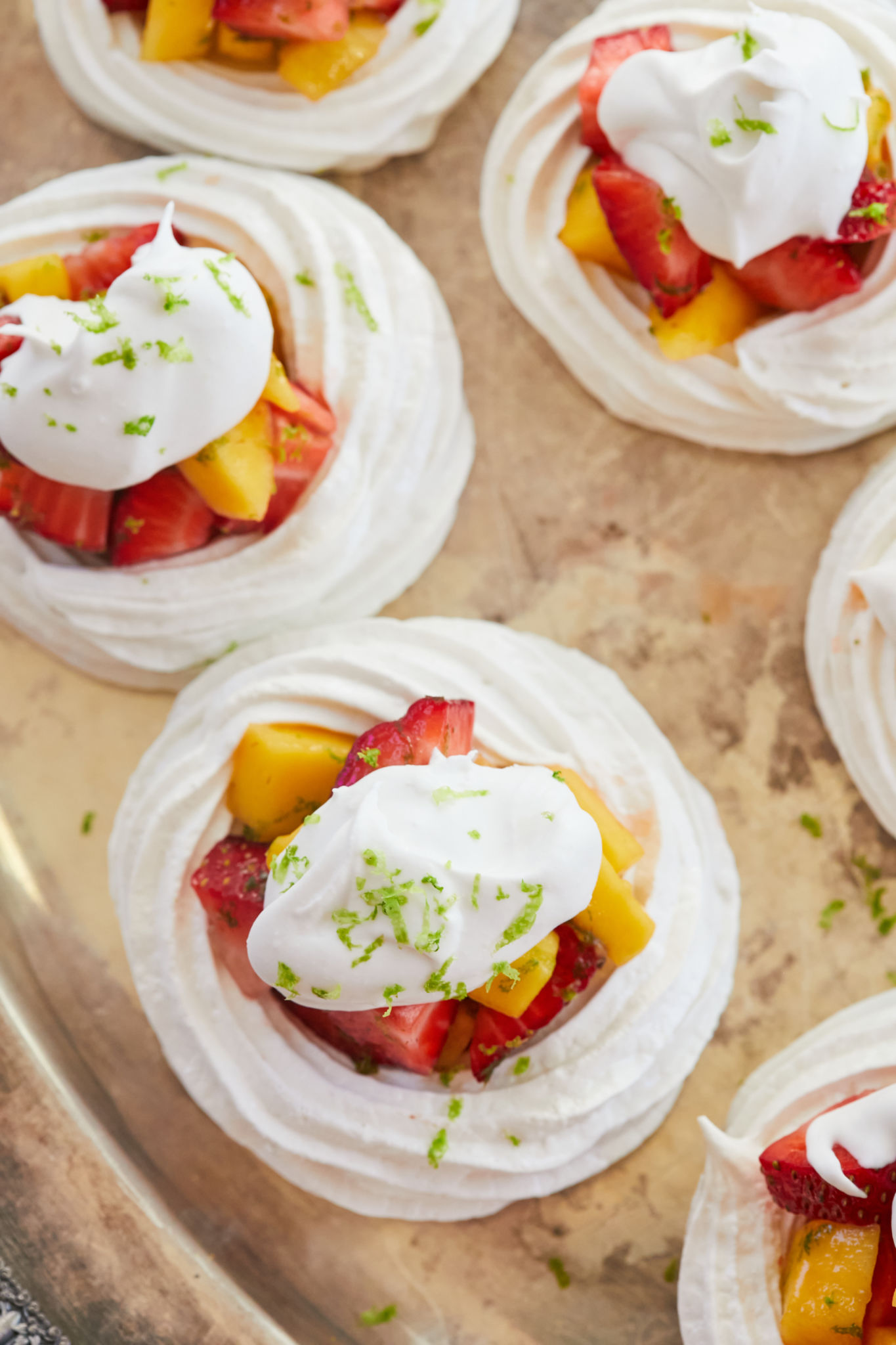 An overhead shot showing Heavenly Meringue Nests on a silver platter with white layers of crispy billowy meringue loaded with irresistible tropical red strawberries and vibrant yellow mangoes, and adorned with velvety coconut cream, and green refreshing lime zest. 