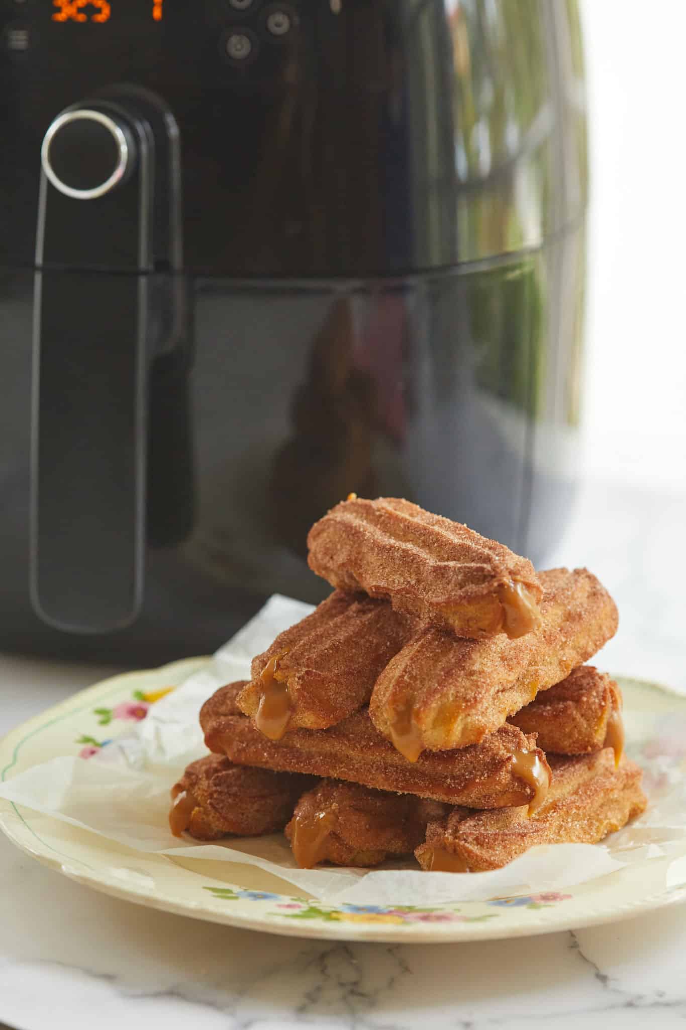 Air fryer churros stacked, filled with dulce de leche, in front of an air fryer.