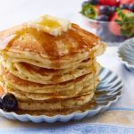 Maple syrup is drizzled over a stack of golden and fluffy pancakes with butter on top and strawberry and blueberries on the side.