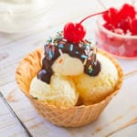 A waffle cone bowl filled with vanilla pudding ice cream, topped with chocolate, sprinkles, and a cherry.