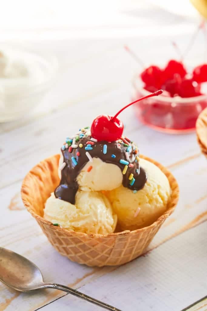 A waffle cone bowl filled with vanilla pudding ice cream, topped with chocolate, sprinkles, and a cherry.