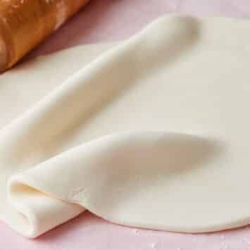 Rolled white fondant draped and folded on a pink surface with a wooden rolling pin, ready for decorating cakes.