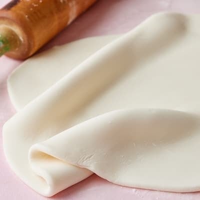 Rolled white fondant draped and folded on a pink surface with a wooden rolling pin, ready for decorating cakes.