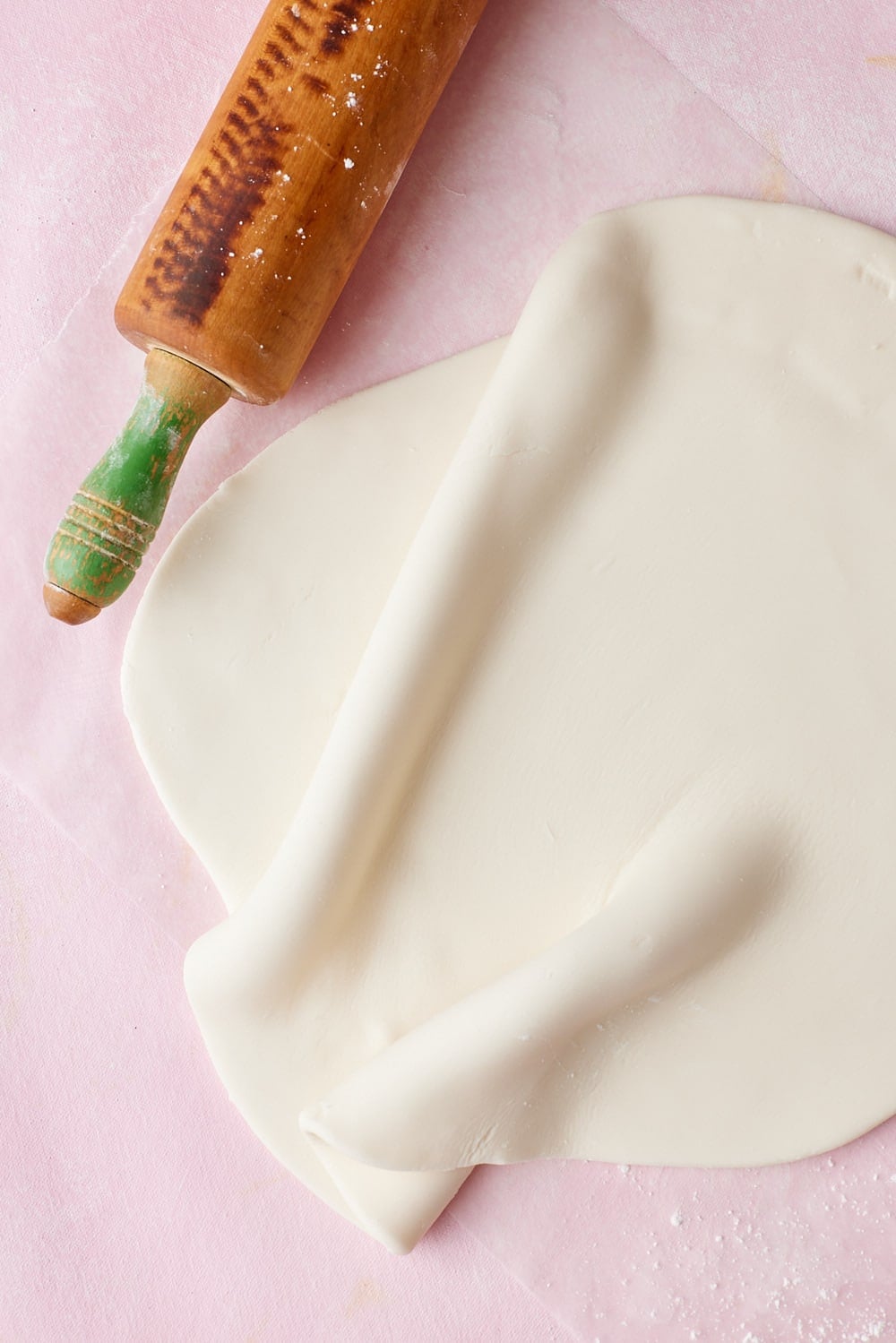 Fondant8 Rolling out smooth white fondant with a wooden rolling pin on a pink work surface, showing the soft, pliable fondant being flattened.