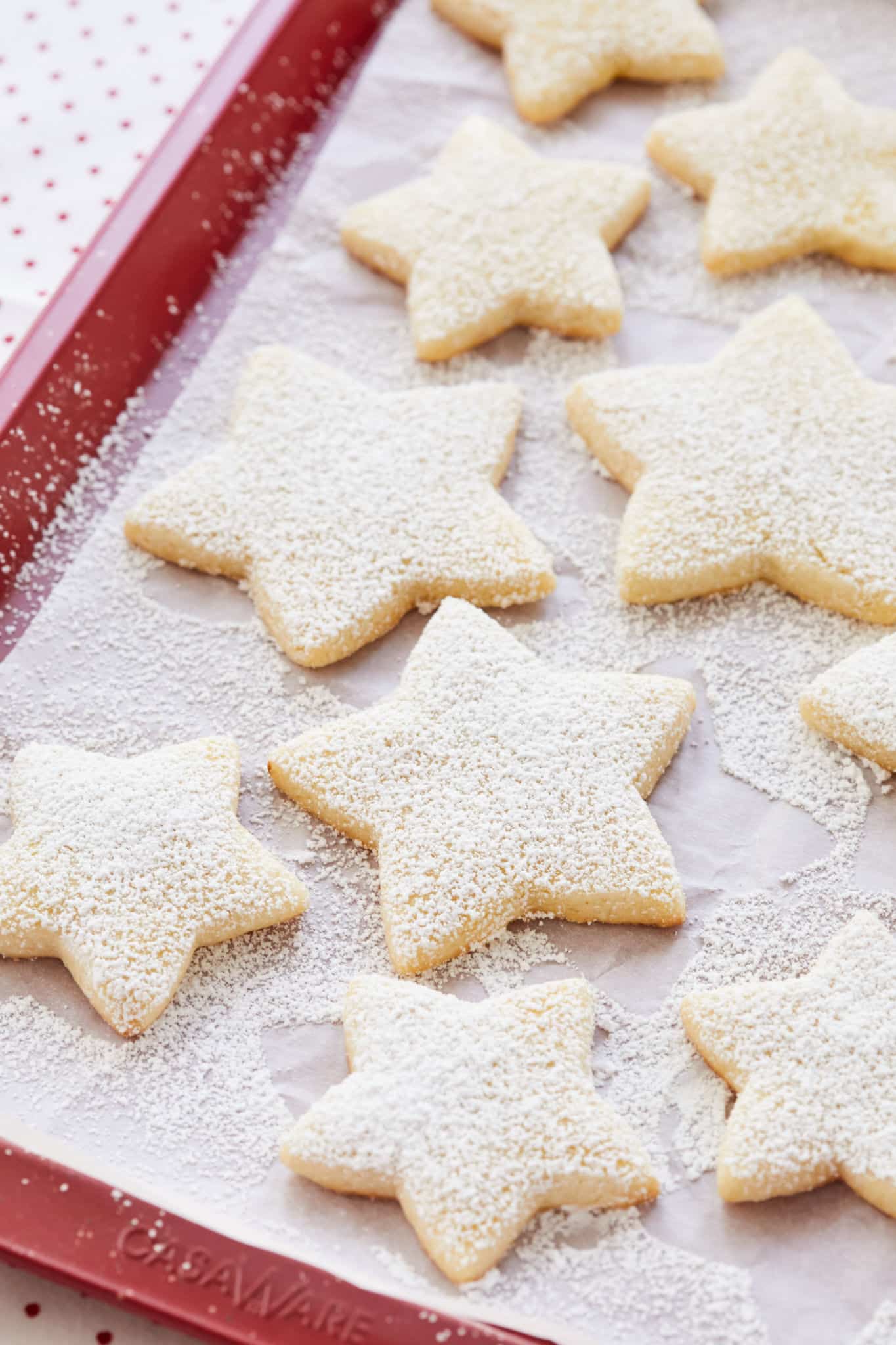 Bredele cookies, cut in the shape of stars, are dusted with powdered sugar on top of a red baking pan layered with parchment paper.