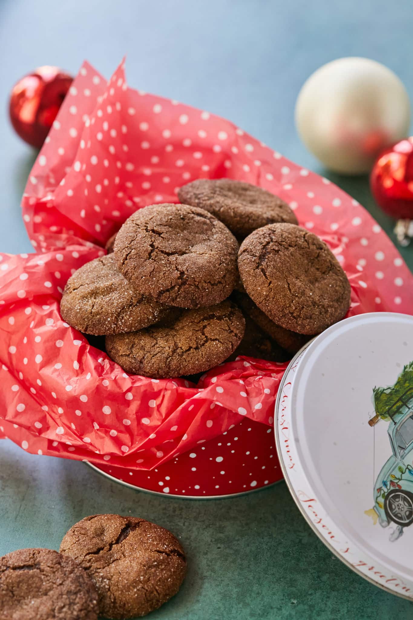 Homemade crispy gingersnaps are served in a Christmas tin with red tissue paper with white polka dots. In the background are Christmas ornaments. 