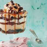 A big glass serving bowl of Chocolate Brownie Trifle with salted caramel dripping down the edge. It's topped with brownie bites.