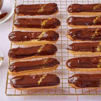 Eclairs glazed with ganache and dusted with edible gold on a wire rack.