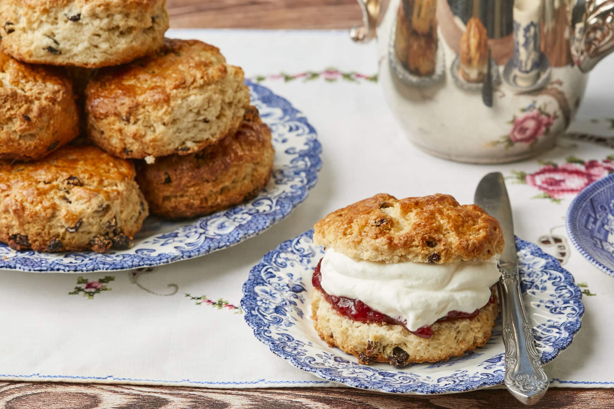 One Irish Scone is served on a floral dessert plate. It is golden brown with a crunchy, crackly exterior and loaded with raisins, stuffed with jam and whipped cream. The rest of the scones are on the big platter.