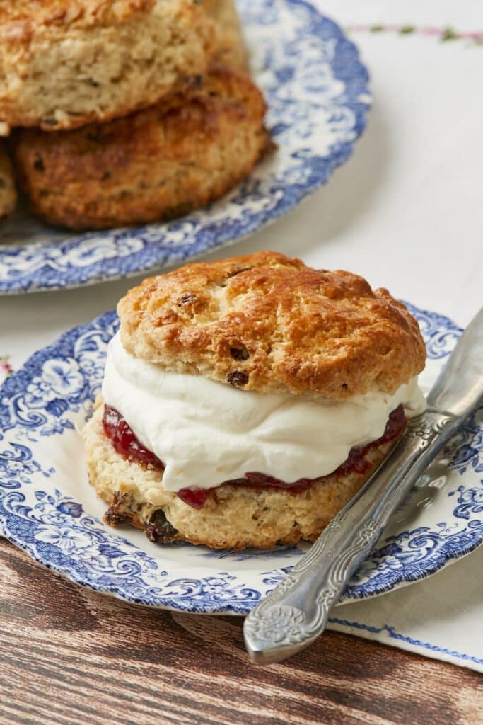 One Irish Scone is served on a floral dessert plate. It is golden brown with a crunchy, crackly exterior and loaded with raisins, stuffed with jam and whipped cream. The rest of the scones are on the big platter.
