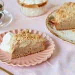 A close-up shot at a slice of Irish Apple Cake served on a dessert plate in the front shows the moist soft cake layer, the sweet apple layer and the crumbly topping. The rest of the cake is on the big platter and a cup of coffee is next to it.