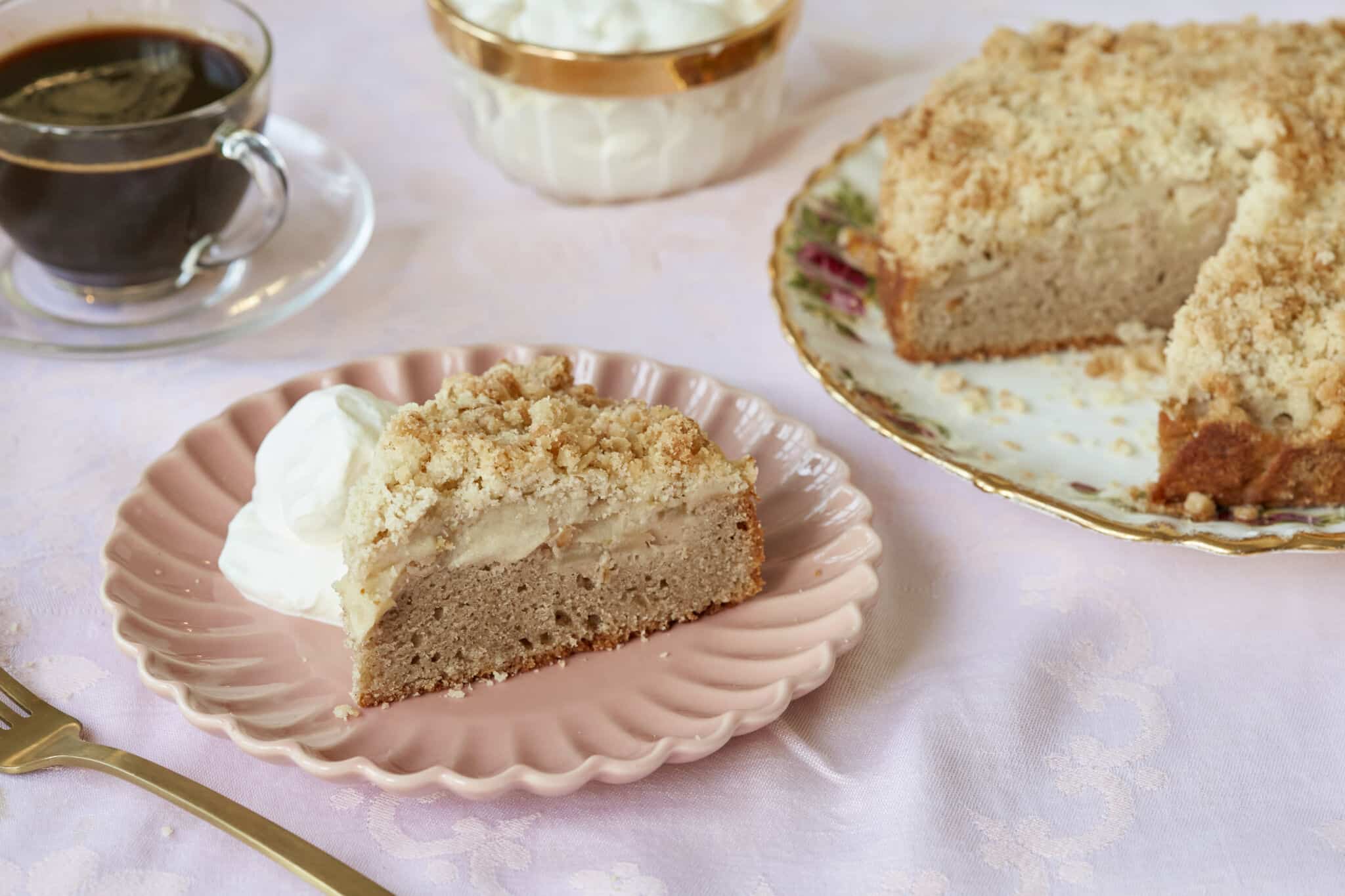 A close-up shot at a slice of Irish Apple Cake served on a dessert plate in the front shows the moist soft cake layer, the sweet apple layer and the crumbly topping. The rest of the cake is on the big platter and a cup of coffee is next to it.