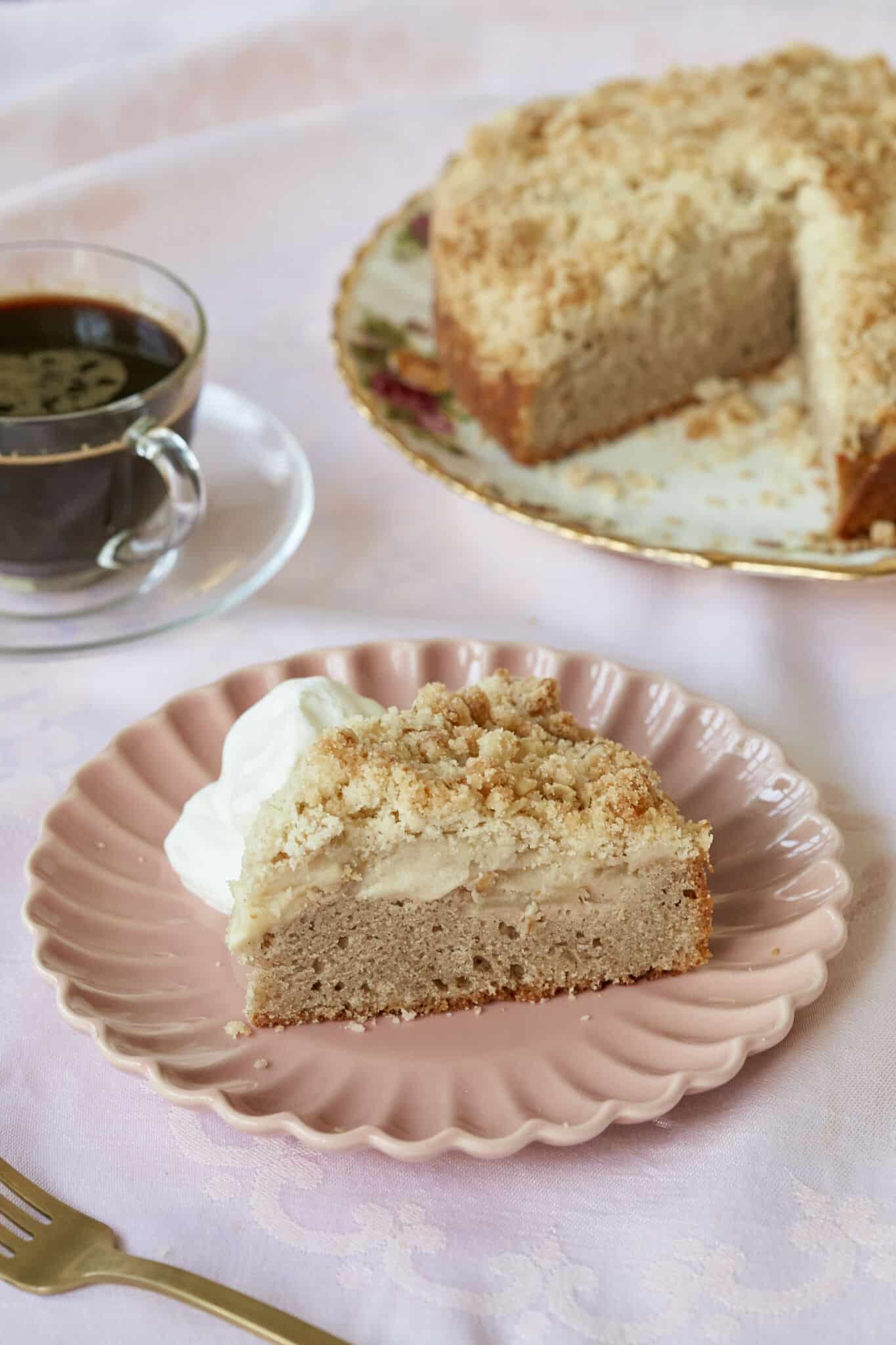 A close-up shot at a slice of Irish Apple Cake served on a dessert plate in the front shows the moist soft cake layer, the sweet apple layer and the crumbly topping. The rest of the cake is on the big platter and a cup of coffee is next to it. 