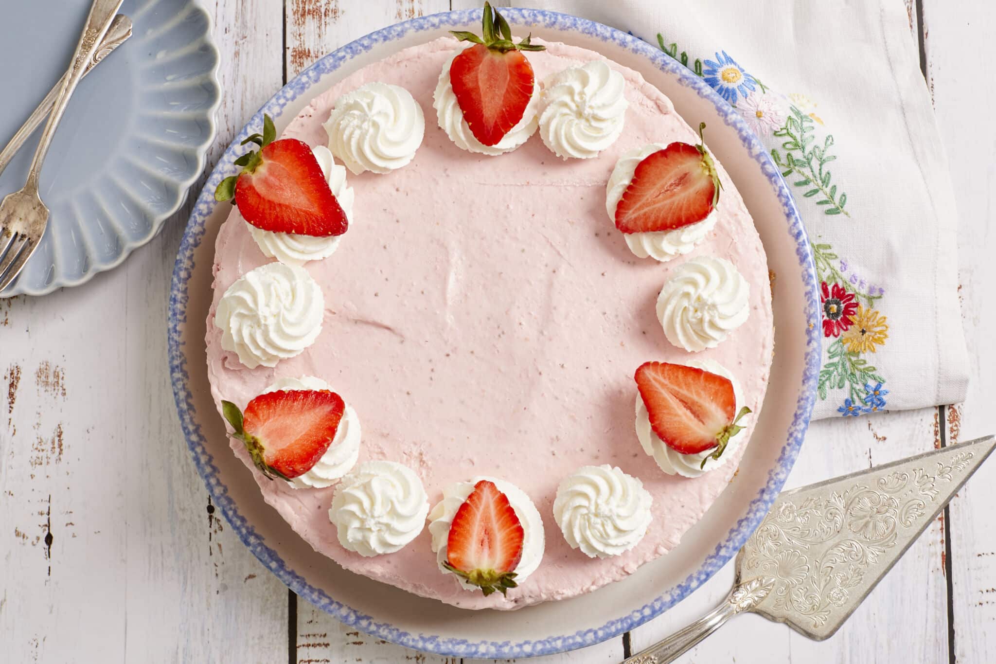 An overhead shot of the whole No-Bake Strawberry Cheesecake on a round platter with bright-light-pink velvety cream cheese filling, topped white whipped rosettes and fresh strawberry halves. A dessert plate with a fork in the upper left corner and a serving knife at the bottom right corner.