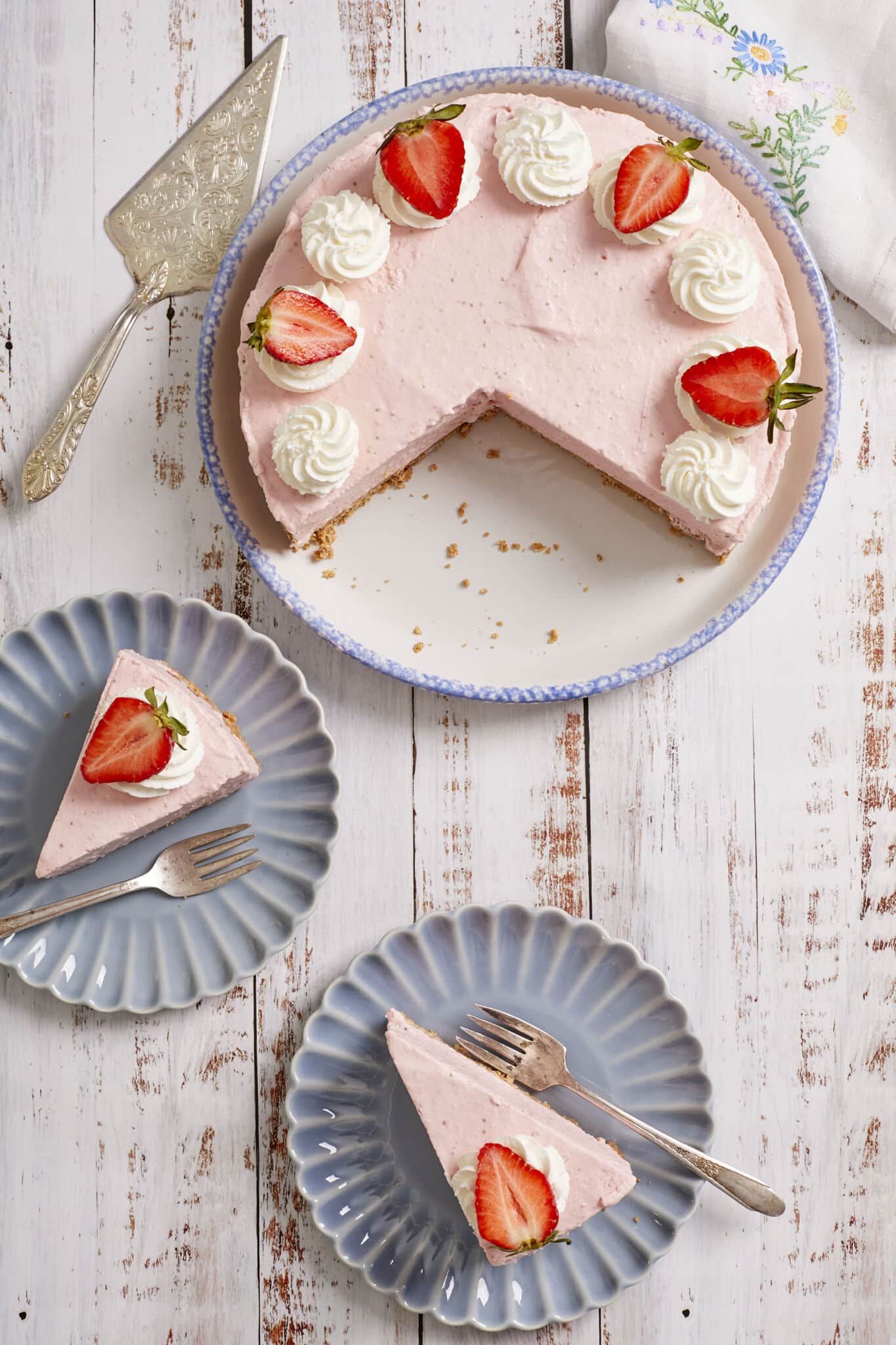 The decadent light-pink No-Bake Strawberry Cheesecake is served on a big platter topped with white whipped cream and red fresh strawberries. Two slices are served individually on two dessert plates with forks. 