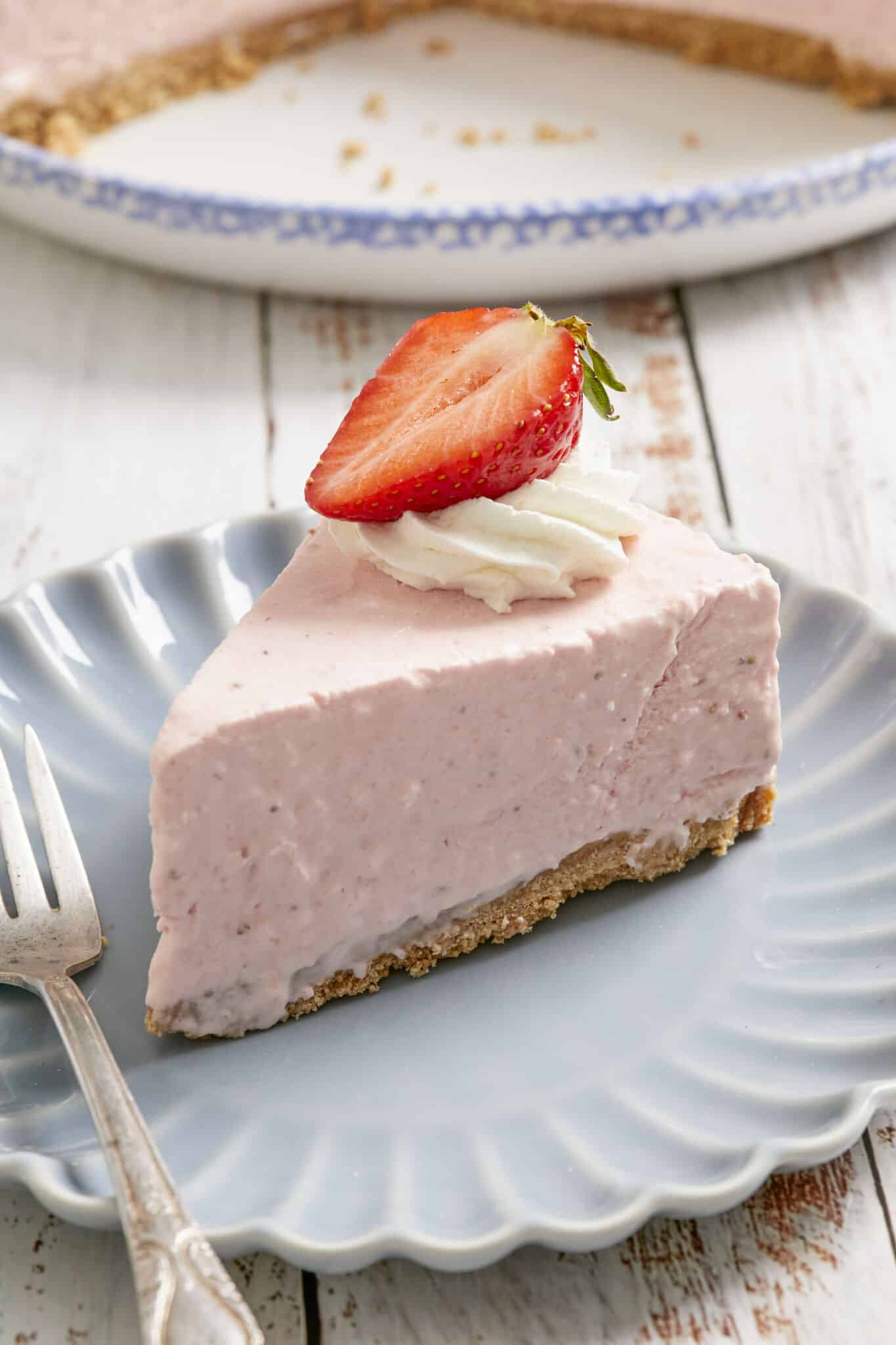 A close shot at a slice of No-Bake Strawberry Cheesecake with cookies crust at the bottom, creamy filling on top, decorated with a whipped cream rosette and half fresh strawberry. 