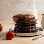 Chocolate pancakes drizzled with chocolate next to strawberries.