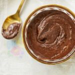 An overhead shot of Homemade Nutella in a glass bowl, with a gold spoon next to it on a floral tablecloth.