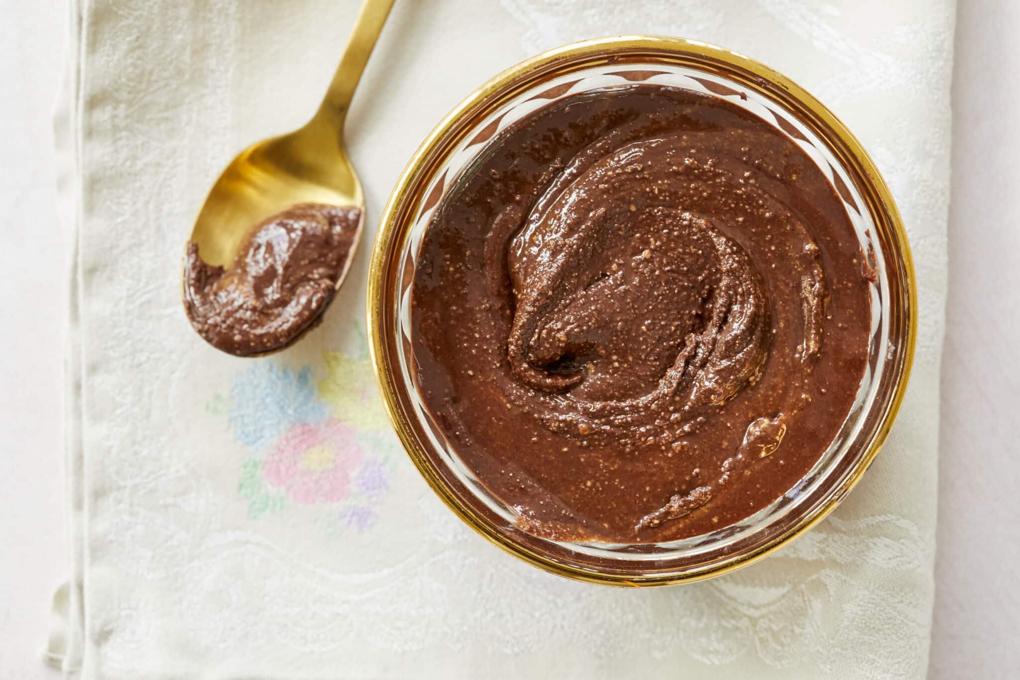 An overhead shot of Homemade Nutella in a glass bowl, with a gold spoon next to it on a floral tablecloth.