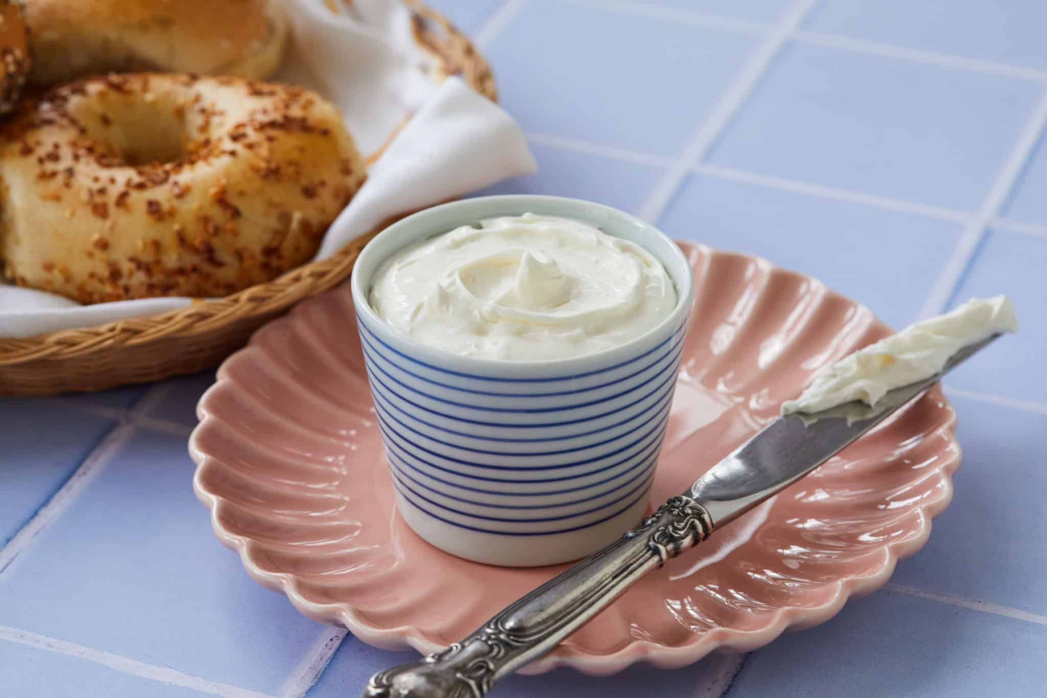 A cup of homemade whipped cream cheese sits on a pink plate, with a butter knife that has been used to swirl it, next to everything bagels.