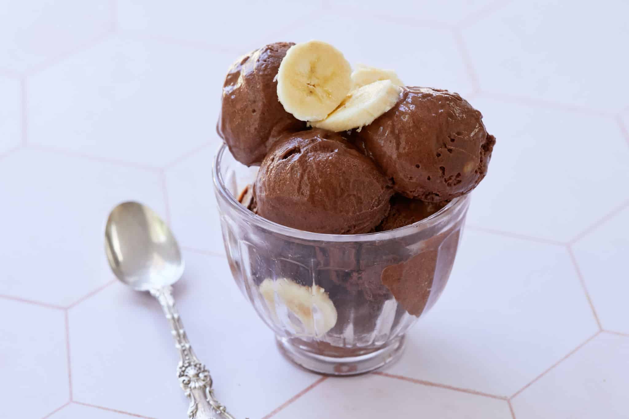 Scoops of chocolate ice cream for breakfast are served in a glass bowl, topped with fresh bananas.