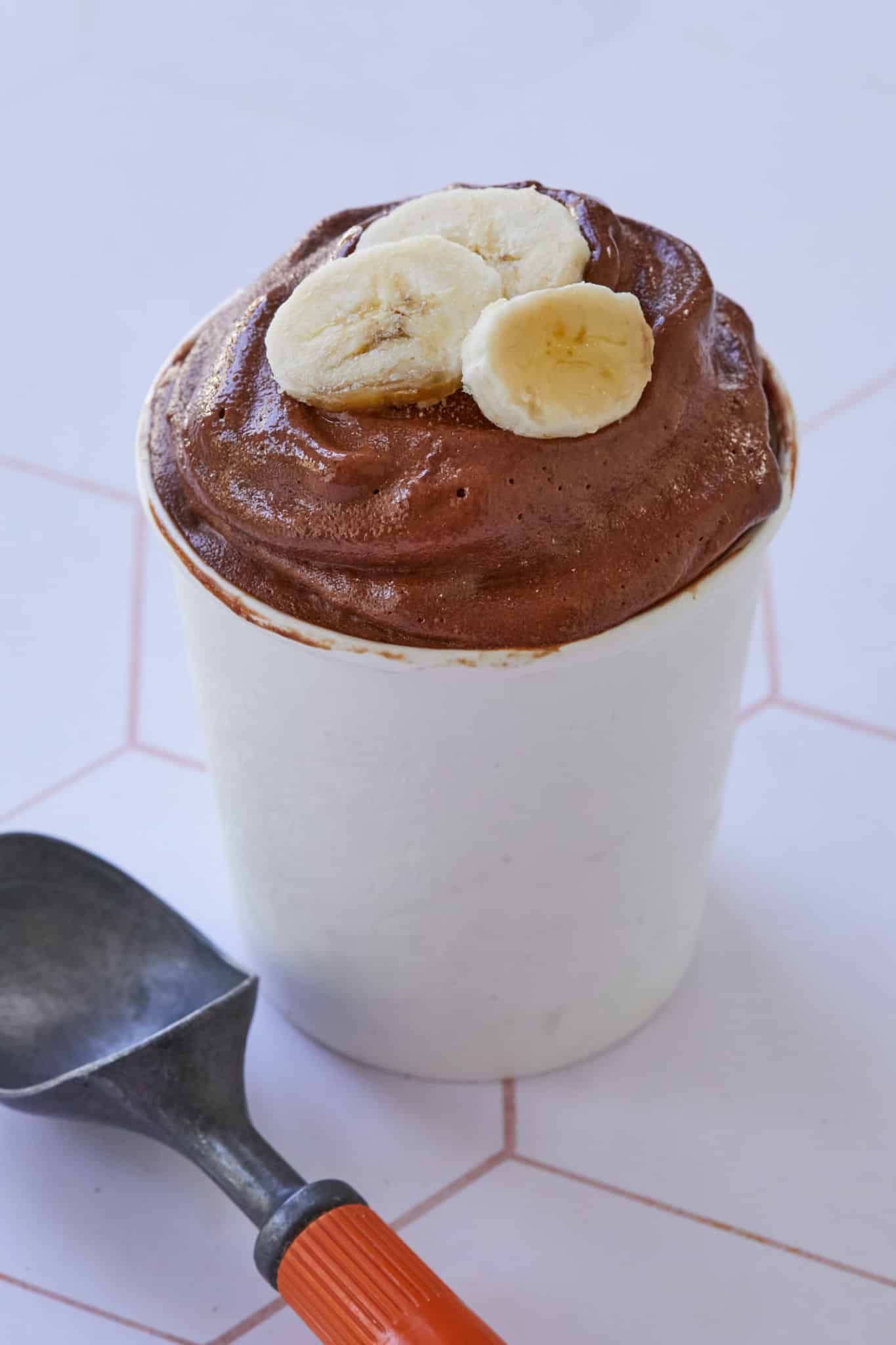 Smooth, cream chocolate ice cream for breakfast is served in a white cup with fresh bananas. This healthy ice cream recipe is made with frozen bananas and dates, perfect for a breakfast treat.