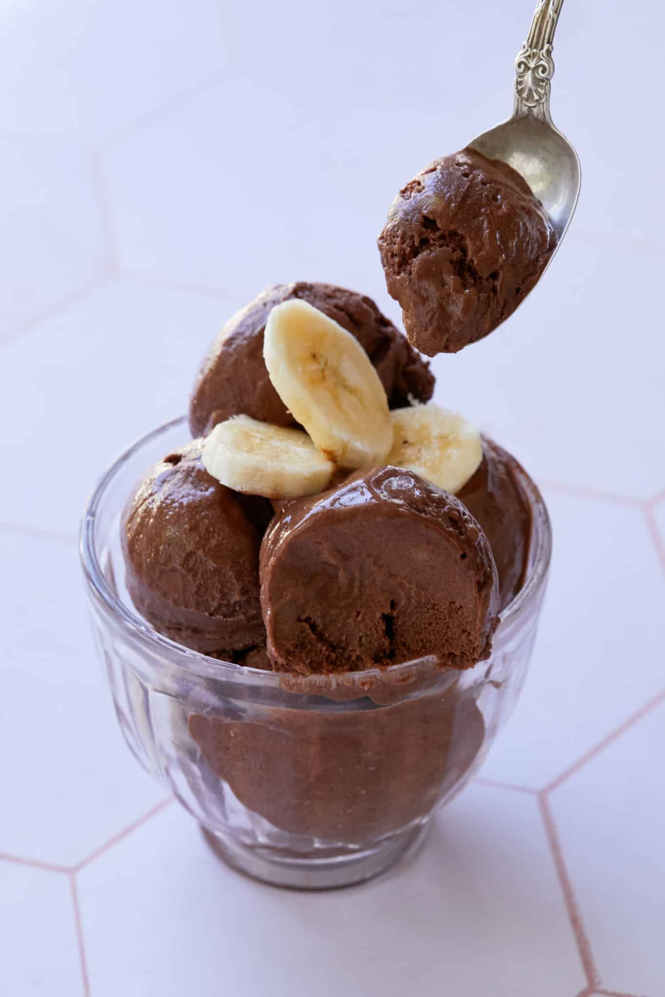 Healthy breakfast ice cream is served in a glass bowl, topped with fresh bananas. A spoonful of the chocolate nice ice is raised above the bowl.