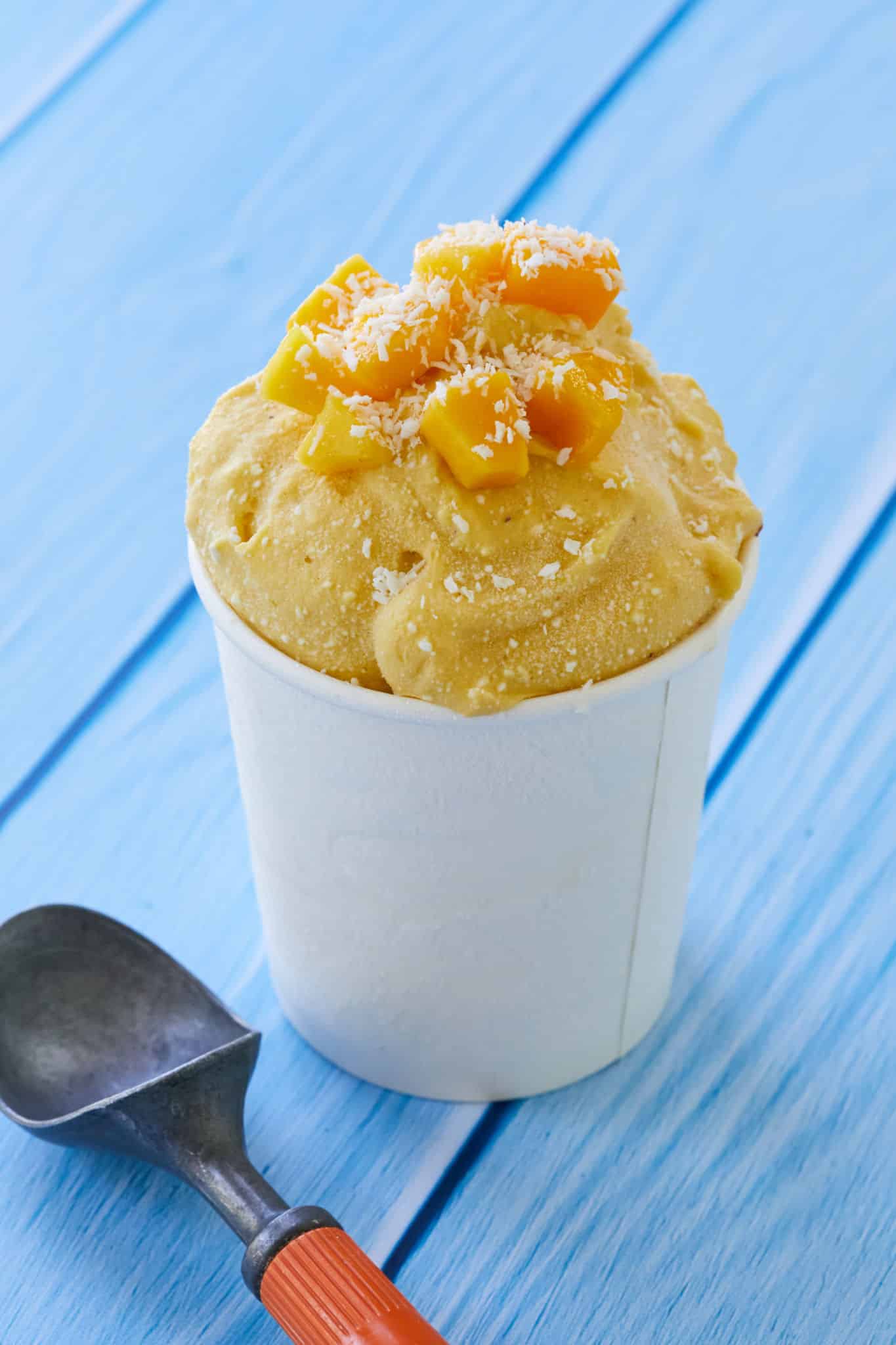 Homemade nice cream is served in a paper cup. The mango coconut ice cream is topped with fresh mango and coconut shavings.