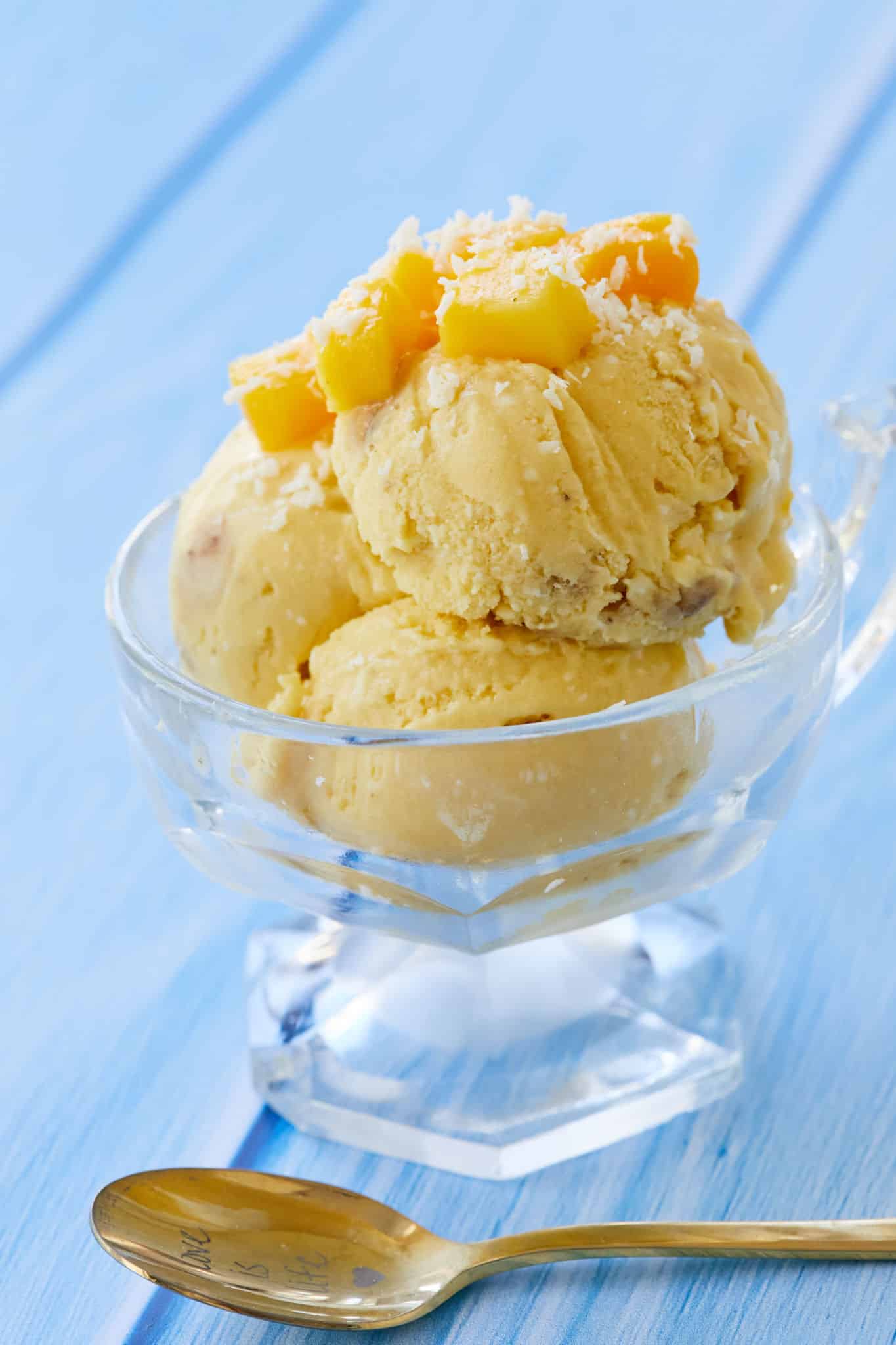 Homemade Mango Coconut Ice Cream For Breakfast is scooped inside of a glass serving cup. The ice cream is topped with mango and coconut shavings. A spoon that reads "Love is Life" rests next to the bowl of ice cream.