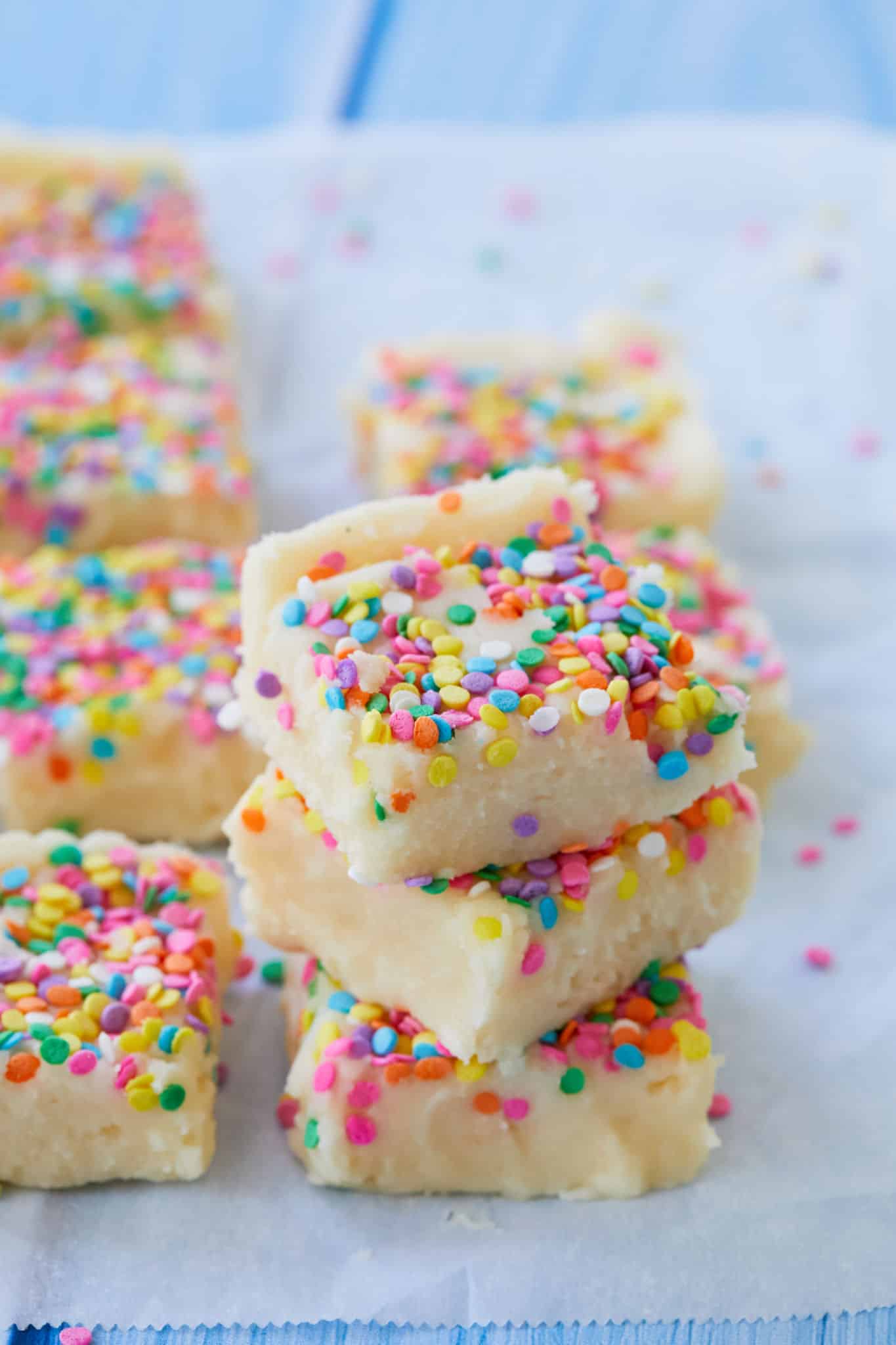 Three squares of Birthday Cake Fudge are stacked on top of each other, with other squares of the white fudge in the background. All of the pieces of homemade old-fashioned fudge are covered in circular rainbow sprinkles.