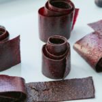 Rolls of homemade blueberry fruit leather line a baking sheet. The fruit leather, made with real blueberries, is a dark purple color.